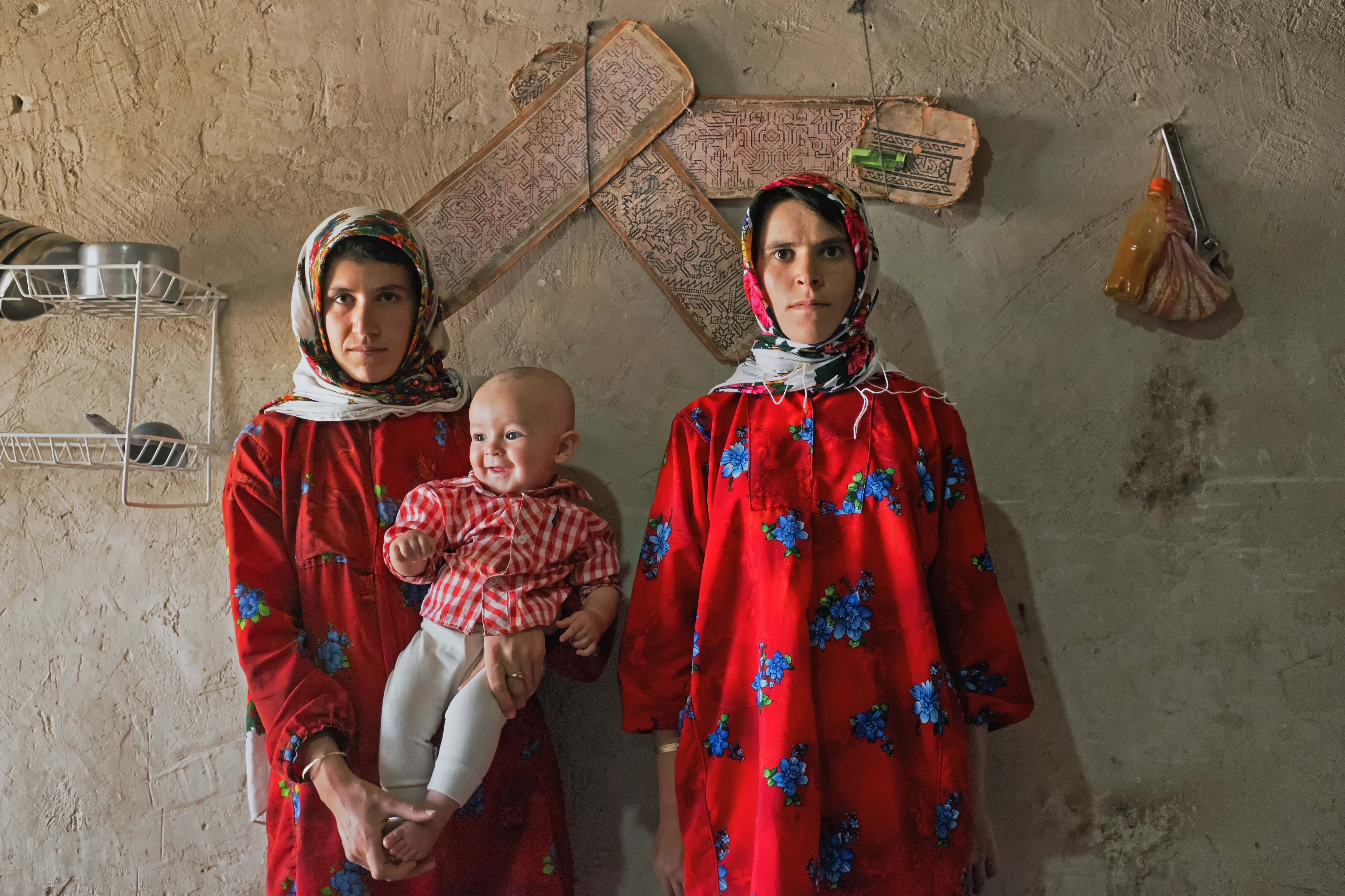 Roghayyeh stands beside Azar her sister-in-law who is holding her son. They weave the carpet in their free time, carpet drafts are seen behind them. Ajabshir, East Azerbaijan, Iran.