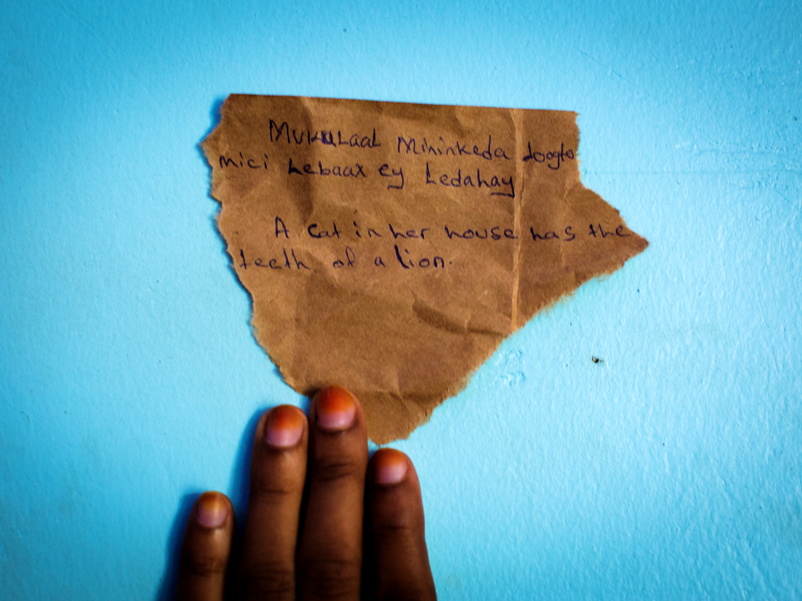 A Somali proverb is written down by one of this project's female protagonists on a small piece of paper and placed on a flower bedsheet in a shared room of female Somali refugees in Kampala's Kisenyi neighbourhood. We cooperated in finding and selecting proverbs to be added to this project.